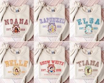Op de afbeelding: Een collectie van zes beige sweaters met ronde hals, elk voorzien van een andere Disney-personageafbeelding. De ontwerpen bevatten namen zoals Moana, Rapunzel, Elsa, Belle, Sneeuwwitje en Tiana, vergezeld van jaartallen en locatienamen.