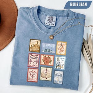 Puede incluir: Una camiseta color azul jean con una cuadrícula de nueve diseños rectangulares de sellos postales. Los gráficos incluyen un castillo etiquetado como Lunathion, una montaña etiquetada como The Night Court, una espada, murciélagos, rosas, un caballete, un ciervo, un libro y alas.