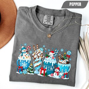 May include: A pepper-colored short-sleeve t-shirt featuring a graphic print of four festive mugs filled with holiday treats, including snowmen, candy canes, Christmas trees, and wrapped gifts, with a Comfort Colors brand label at the neck.