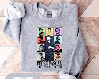 Könnte beinhalten: Ein sportgraues Sweatshirt mit Rundhalsausschnitt und einem Grafikdruck von Pedro Pascal in verschiedenen Rollen, angeordnet in einem Raster. Unter dem Raster steht der Text PEDRO PASCAL THE ERAS TOUR. Das Kleidungsstück hat lange Ärmel sowie gerippte Bündchen.