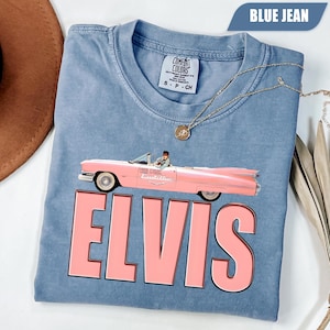 May include: A blue jean colored t-shirt featuring a graphic of a pink Cadillac convertible with a person inside. Below the car, the word ELVIS is printed in large, bold, light pink capital letters. The shirt has a Comfort Colors brand label.