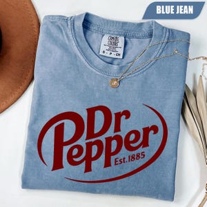 Puede incluir: Una camiseta de color azul vaquero con un gran logotipo rojo de Dr Pepper y el texto Est. 1885. La prenda tiene cuello redondo y una etiqueta de la marca Comfort Colors en el interior del cuello. Una fina cadena dorada con un peque&ntilde;o colgante circular descansa sobre ella.