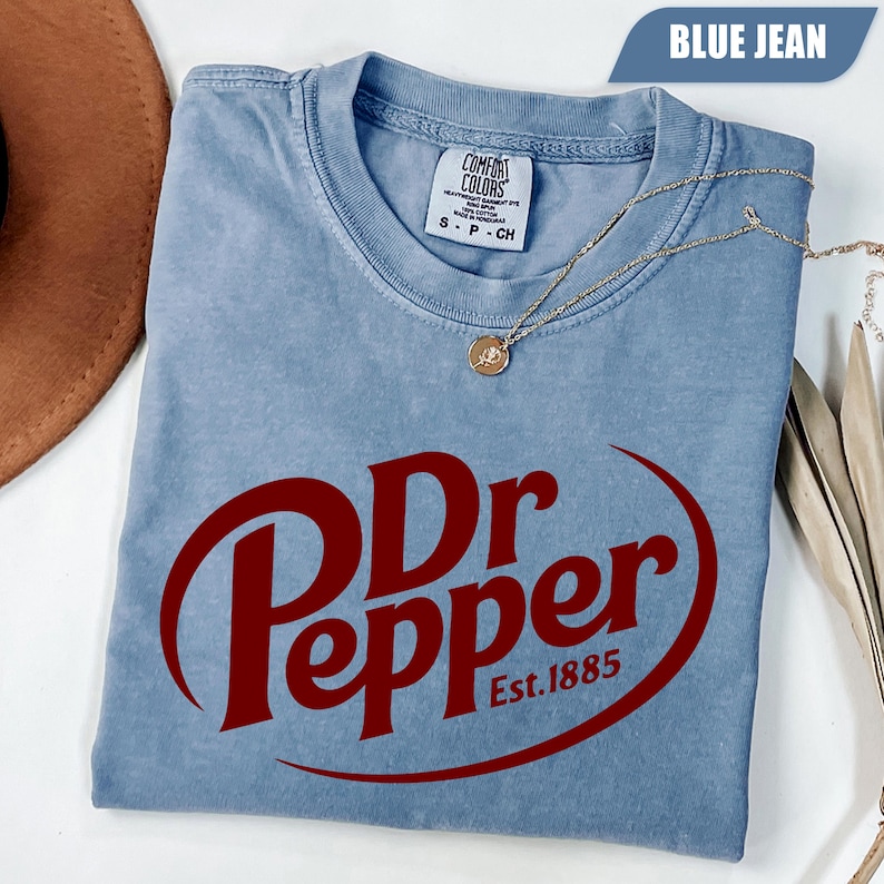 Puede incluir: Una camiseta de color azul vaquero con un gran logotipo rojo de Dr Pepper y el texto Est. 1885. La prenda tiene cuello redondo y una etiqueta de la marca Comfort Colors en el interior del cuello. Una fina cadena dorada con un peque&ntilde;o colgante circular descansa sobre ella.