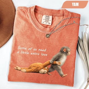Puede incluir: Una camiseta de manga corta color coral con un gráfico de un mono gris tirando de un mono de peluche marrón sobre la tela. El texto "Some of us need a little extra love" está impreso en blanco sobre el gráfico. Tiene cuello redondo.