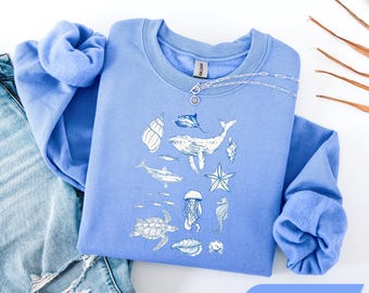 Könnte beinhalten: Ein Carolina-blaues Sweatshirt mit Rundhalsausschnitt und einem weißen Grafikdruck verschiedener Meeresbewohner, darunter ein Wal, eine Meeresschildkröte, eine Qualle, ein Seepferdchen, ein Seestern, ein Hai, ein Rochen und Muscheln.