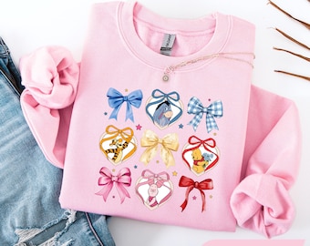 Könnte beinhalten: Ein hellrosa Sweatshirt mit Rundhalsausschnitt und einem Grafikdruck aus verschiedenen Schleifen und herzförmigen Rahmen, die Illustrationen von I-Aah, Tigger, Ferkel und Winnie Puuh enthalten. Das Sweatshirt hat lange Ärmel.