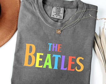 Könnte beinhalten: Ein anthrazitfarbenes Kurzarm-T-Shirt mit Rundhalsausschnitt. Das T-Shirt zeigt den Schriftzug THE BEATLES in einer mehrfarbigen Regenbogen-Schriftart auf der Brust. Eine dünne goldfarbene Kette mit einem kleinen runden Anhänger liegt auf dem Kragen.