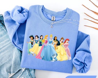 Könnte beinhalten: Ein Carolina-blaues Sweatshirt mit Rundhalsausschnitt und einem Grafikdruck von neun animierten Prinzessinnen, die in einer Reihe stehen. Das Sweatshirt hat lange Ärmel und einen gerippten Kragen. Eine silberfarbene Kette mit einem kleinen runden Anhänger liegt am Halsausschnitt.