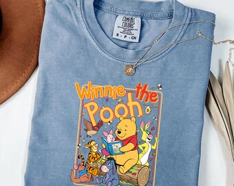 Könnte beinhalten: Ein blaues T-Shirt in Jeansoptik mit einem Grafikdruck von Winnie Puuh und seinen Freunden, die ein Buch lesen. Die Figuren sind Tigger, Ferkel, I-Aah, Rabbit und Ruh. Der Text lautet Winnie the Pooh. Das Shirt hat ein Comfort Colors Etikett.