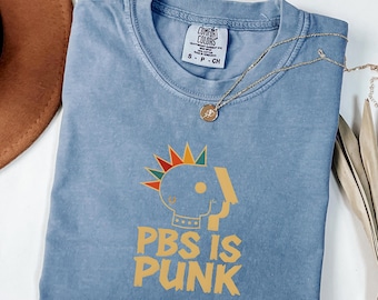 Könnte beinhalten: Ein blaues T-Shirt in Jeansoptik mit einer Grafik eines stilisierten Kopfes mit einem mehrfarbigen Irokesenschnitt und dem gelben Schriftzug PBS IS PUNK. Das Shirt hat einen Rundhalsausschnitt und ein Comfort Colors Etikett am Kragen.