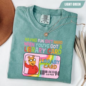 May include: A light green t-shirt featuring the text "HAVING FUN ISN'T HARD WHEN YOU'VE GOT A LIBRARY CARD" in colorful letters. Below the text is a graphic of a library card with a cartoon aardvark and the words "LIBRARY CARD" and "2101908".