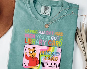 Könnte beinhalten: Ein hellgrünes T-Shirt mit dem Text "HAVING FUN ISN'T HARD WHEN YOU'VE GOT A LIBRARY CARD" in bunten Buchstaben. Unter dem Text befindet sich eine Grafik eines Bibliotheksausweises mit einem Erdferkel und den Wörtern "LIBRARY CARD" und "2101908".