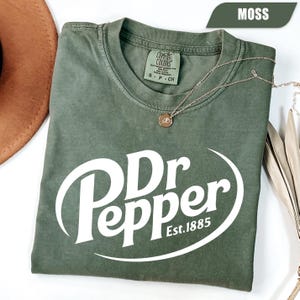 Puede incluir: Una camiseta de manga corta color verde musgo con el logotipo blanco de Dr Pepper y el texto Est. 1885 impreso en la parte delantera. La prenda tiene un cuello redondo con una etiqueta de la marca Comfort Colors y una cadena dorada fina.