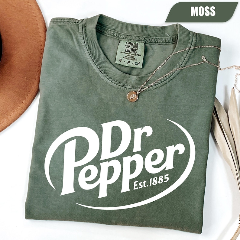 Puede incluir: Una camiseta de manga corta color verde musgo con el logotipo blanco de Dr Pepper y el texto Est. 1885 impreso en la parte delantera. La prenda tiene un cuello redondo con una etiqueta de la marca Comfort Colors y una cadena dorada fina.