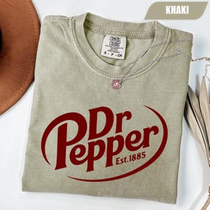 Puede incluir: Una camiseta de manga corta color caqui con el logotipo rojo oscuro de Dr Pepper y el texto Est. 1885. La prenda tiene cuello redondo y una etiqueta de la marca Comfort Colors en el interior. Un collar de cadena plateada descansa sobre la tela.