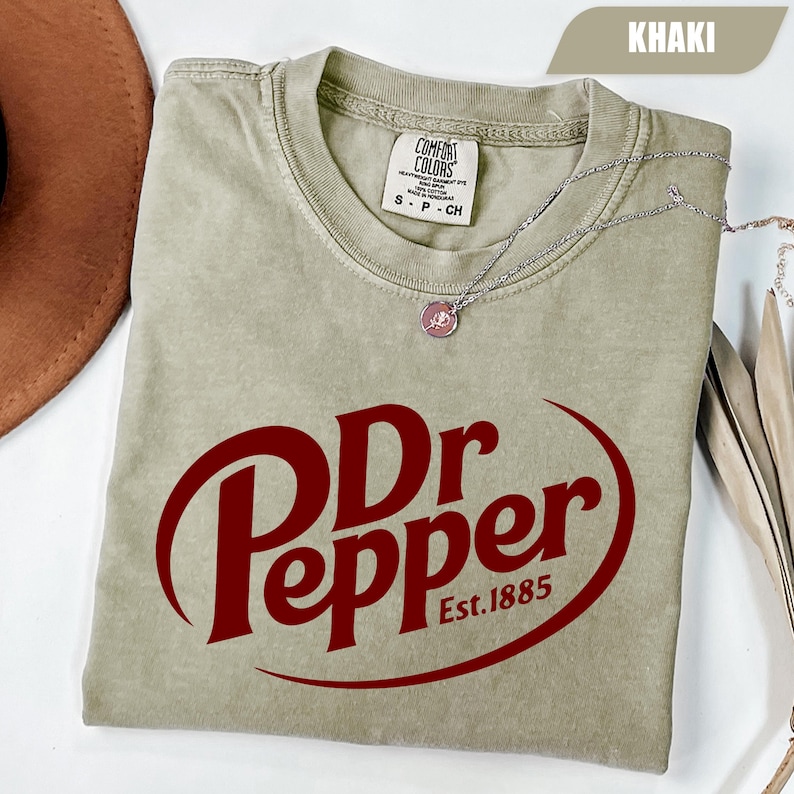 Puede incluir: Una camiseta de manga corta color caqui con el logotipo rojo oscuro de Dr Pepper y el texto Est. 1885. La prenda tiene cuello redondo y una etiqueta de la marca Comfort Colors en el interior. Un collar de cadena plateada descansa sobre la tela.