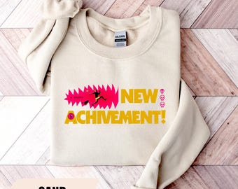 Op de afbeelding: Een zandkleurige sweater met ronde hals en een grafische print met de tekst "NEW ACHIVEMENT!" in het geel. Het ontwerp bevat een roze grillige vorm, een klein figuurtje en drie kleine roze schedelpictogrammen. Voorzien van Gildan-label.