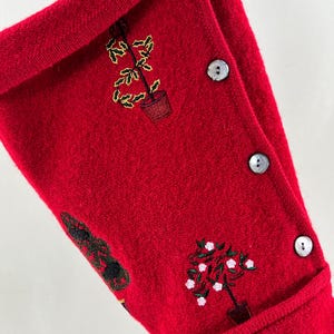 May include: Red knitted Christmas stocking with embroidered green and yellow plants and white flowers. The stocking has four white buttons down the side.
