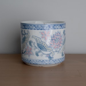 May include: A cylindrical ceramic cup featuring a white base with a repeating blue and pink floral and butterfly pattern. The top and bottom edges are decorated with a detailed blue geometric border. The cup has a smooth, glossy finish.