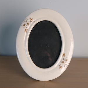 May include: An oval-shaped ceramic picture frame with a cream-colored, crackled glaze finish. The frame features delicate, hand-painted floral motifs in shades of brown and white, positioned on the upper left and lower right sides of the border.