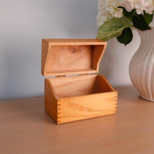May include: A rectangular wooden box with a light brown natural wood grain finish and a hinged lid that opens upward. The box features visible finger joint construction at the corners and a smooth interior surface, showcasing the organic texture of the wood.