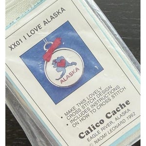 May include: A cross stitch kit titled XX01 I LOVE ALASKA by Calico Cache, featuring a small circular hoop with a blue map of Alaska and a red heart. The packaging includes instructions, a red ribbon, and the text Naomi Leonard 1992.