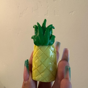 May include: A small, three-dimensional pineapple figurine featuring a bright yellow, textured body with a diamond-shaped grid pattern and a vibrant green, leafy top. The object has a smooth, glossy finish and is held upright in a vertical orientation.