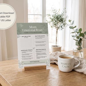 Peut inclure: Une feuille imprimée intitulée Mom Emotional Reset avec des sections pour la réflexion quotidienne, les moments de gratitude, les exercices de pleine conscience et de respiration, sur un support en bois près d'une tasse blanche.