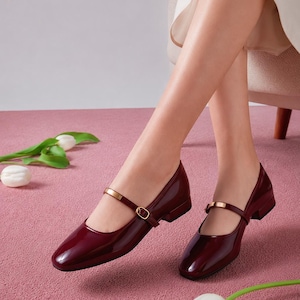 Könnte beinhalten: Ein Paar dunkelburgunderrote Mary-Jane-Schuhe aus Lackleder mit niedrigem Blockabsatz und einem dünnen Riemen über dem Spann, der mit einer goldenen Metallschnalle und einem Zierelement versehen ist. Die Schuhe haben eine runde Zehenpartie.