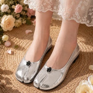 May include: A pair of metallic silver split-toe flats featuring a textured finish and a small black decorative knot detail at the center of the vamp. The shoes have a low profile and a distinct cleft design separating the big toe from the rest of the foot.
