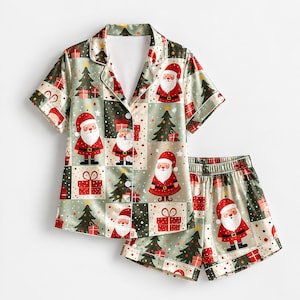 May include: A short-sleeve button-down pajama shirt and matching elastic-waist shorts set in a light sage green color. The fabric features a repeating grid pattern of Santa Claus figures, decorated Christmas trees, and red gift boxes with bows.