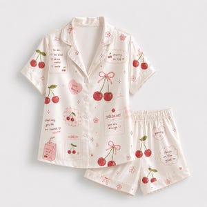 May include: A cream-colored pajama set featuring a short-sleeved button-down shirt and matching shorts. The fabric is decorated with red cherry illustrations, pink bows, and text boxes containing phrases like to do, cherry on top, and note to self.