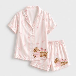 May include: A light pink pajama set featuring a short-sleeved button-down shirt and matching shorts. The fabric has vertical white stripes, small pink bow patterns, and illustrations of brown dachshunds wearing striped shirts with pink flowers.