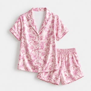 May include: A light pink pajama set consisting of a short-sleeved button-down shirt with a notched collar and matching elastic-waist shorts. The fabric features a repeating pattern of dark pink floral vines and stylized animal figures.