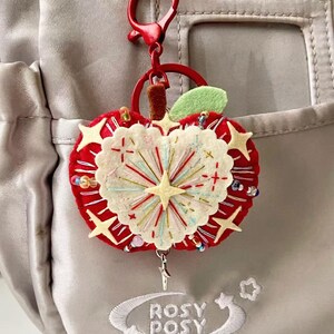 May include: A red felt apple keychain featuring a cream-colored heart-shaped center with embroidered starburst patterns, colorful beads, and a green leaf. The keychain is attached to a red plastic lobster clasp and ring, hanging from a light gray bag.