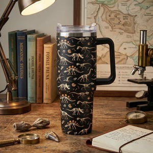 May include: A black stainless steel tumbler with a handle and a clear lid, featuring a repeating pattern of gold-colored dinosaur skeletons and fossils. The tall, cylindrical drinkware has a wide base and a sturdy side handle for gripping.