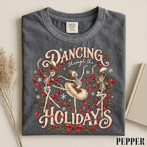 May include: A charcoal gray t-shirt featuring a graphic of three skeletons dancing. The text reads DANCING through the HOLIDAYS. The skeletons wear festive attire including a tutu and hats, surrounded by red ribbons, holly, and snowflakes.