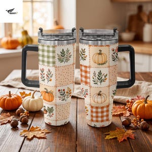 May include: Two stainless steel tumblers with black handles and clear lids, featuring a patchwork design of pumpkins, autumn leaves, and checkered patterns in shades of orange, green, and cream, set against a rustic wooden table surface.
