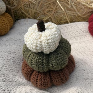 May include: A stack of three crocheted pumpkins in varying sizes and colors. The top pumpkin is cream with a dark brown stem, the middle pumpkin is olive green, and the bottom pumpkin is brown. Each pumpkin features a ribbed, textured yarn surface.