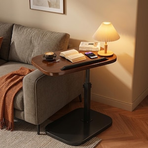 May include: A height-adjustable side table with a dark wood-grain finish top and a black metal base. The table features a black edge guard, a pleated lamp, a book, a smartphone, and a cup of coffee on a saucer, positioned next to a gray sofa.