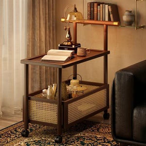 May include: A two-tier rolling trolley with a dark brown wooden frame and light-coloured woven cane side panels. The top shelf holds an open book, a textured ceramic mug, and a brass-toned lamp. The bottom shelf contains various glass bottles and jars.