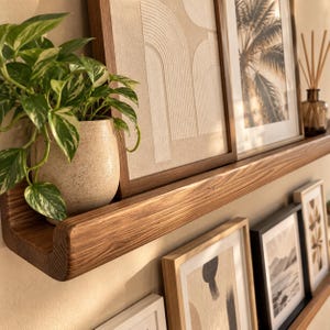 May include: A wooden floating shelf holding a speckled ceramic pot with a green and white variegated pothos plant, a framed beige textured art piece, a framed palm tree print, and a glass bottle with reed diffuser sticks inside.