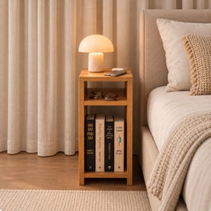 May include: A light oak wood bedside table with an open shelf design. The top surface holds a mushroom-shaped lamp and a smartphone. The middle shelf contains a pair of spectacles. The bottom shelf holds four books titled Rich Dad Poor Dad, Atomic Habits, Think Again, and another book.