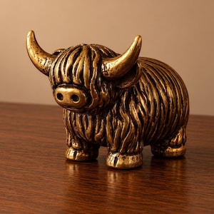 May include: A small, metallic gold-toned figurine of a Highland cow. The sculpture features a textured, shaggy coat, prominent curved horns, and a rounded snout. The surface has a polished, reflective finish with darker recessed areas.
