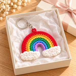 May include: A crocheted rainbow keyring featuring concentric arcs of red, orange, yellow, green, blue, and purple yarn, with white textured clouds at each end. The keyring includes a silver-toned metal split ring attached to the top centre.