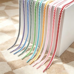 Op de afbeelding: Een collectie platte veters met een herhalend dambordpatroon in diverse kleuren, waaronder zwart, paars, blauw, groen, turquoise, oranje, roze en rood. Elke veter heeft een witte basis met gekleurde vierkantjes en plastic uiteinden.