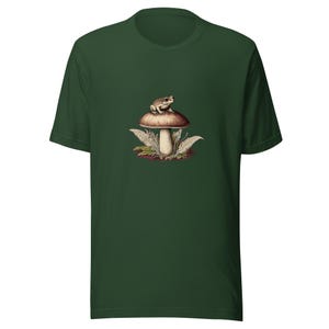 Könnte beinhalten: Ein dunkelwaldgrünes T-Shirt mit einer zentralen grafischen Illustration einer kleinen, hellen Kröte, die auf einem großen Pilz mit braunem Hut sitzt, umgeben von zartem, hellgrünem, farnartigem Blattwerk am Fuß des Pilzstiels.