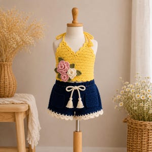 Könnte beinhalten: Ein zweiteiliges Häkelset bestehend aus einem leuchtend gelben Neckholder-Top mit dreidimensionalen rosa und weißen Blumenapplikationen sowie einer dunkelblauen Shorts mit cremefarbener Kordel mit Quasten und einem cremefarbenen Saum.