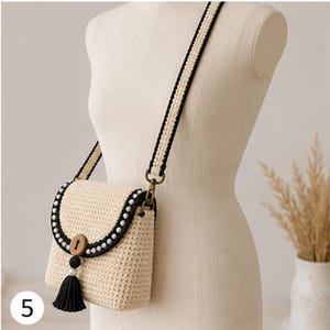 Könnte beinhalten: Eine beige gewebte Umhängetasche mit schwarzem Rand, einem runden Holzknopfverschluss und einer schwarzen Quaste. Die Tasche hat einen langen, verstellbaren Riemen mit einem schwarz-beigen Webmuster, akzentuiert durch kleine weiße Perlen am Rand.