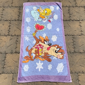 May include: A purple beach towel with a cartoon image of a coyote and a bird. The coyote is holding a gift box and the bird is holding an arrow. The background is a light blue with white snowflakes.